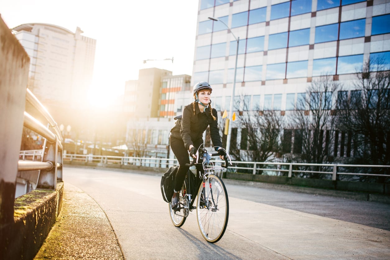 A smiling young woman commuting in an urban city environment on her street bicycle, waterproof panniers on her bike rack. She rides up a ramp from downtown Portland, Oregon, that leads up to the Burnside bridge. Colorful sky from the setting sun. Horizontal image with copy space. A depiction of young adult / hipster lifestyle in the city.