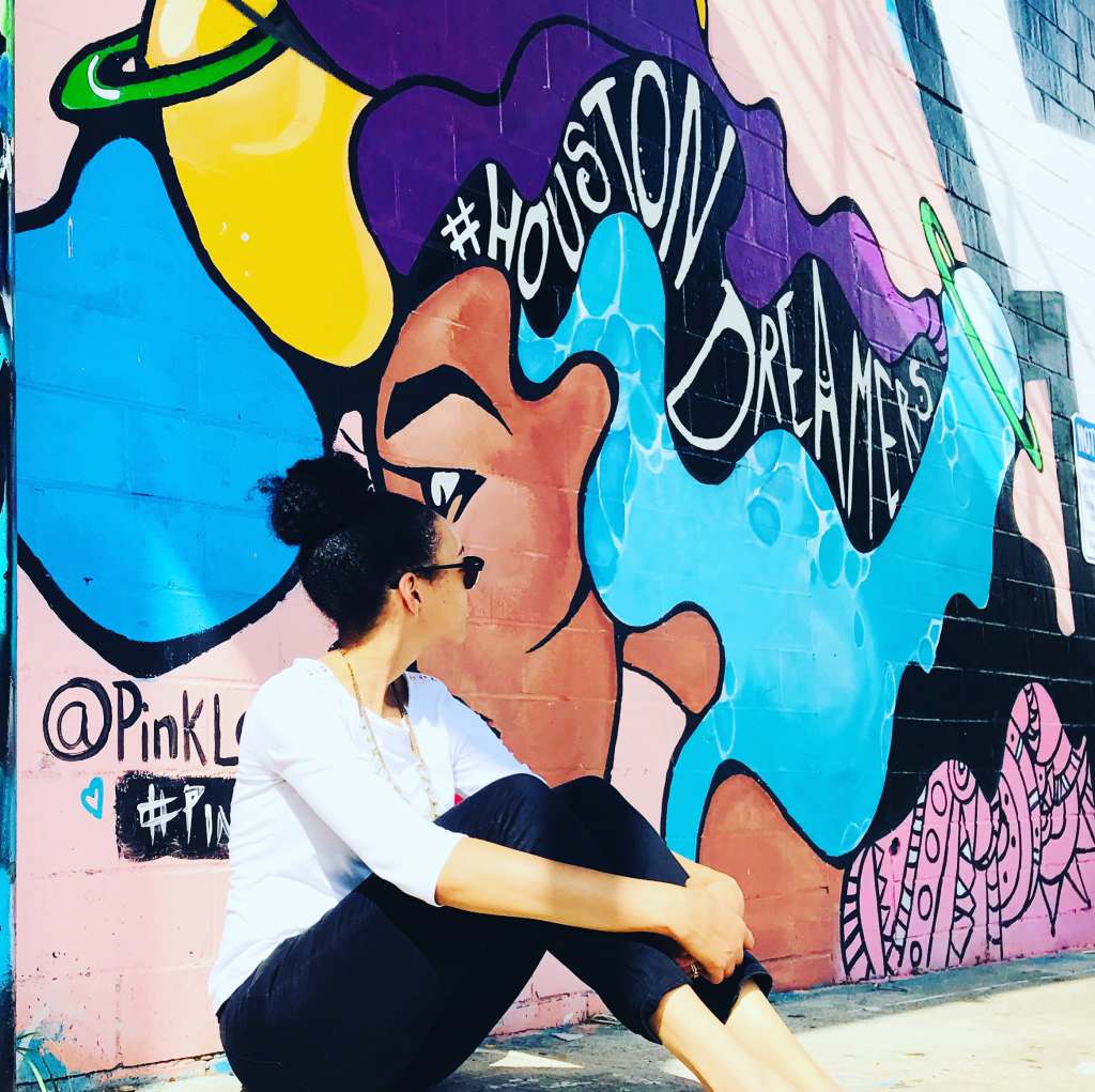 Cities with Incredible Black American Culture A woman sitting in front of a vibrant mural located on Leeland Street in Houston, Texas. The mural is created by an artist known as @PinkLomein.