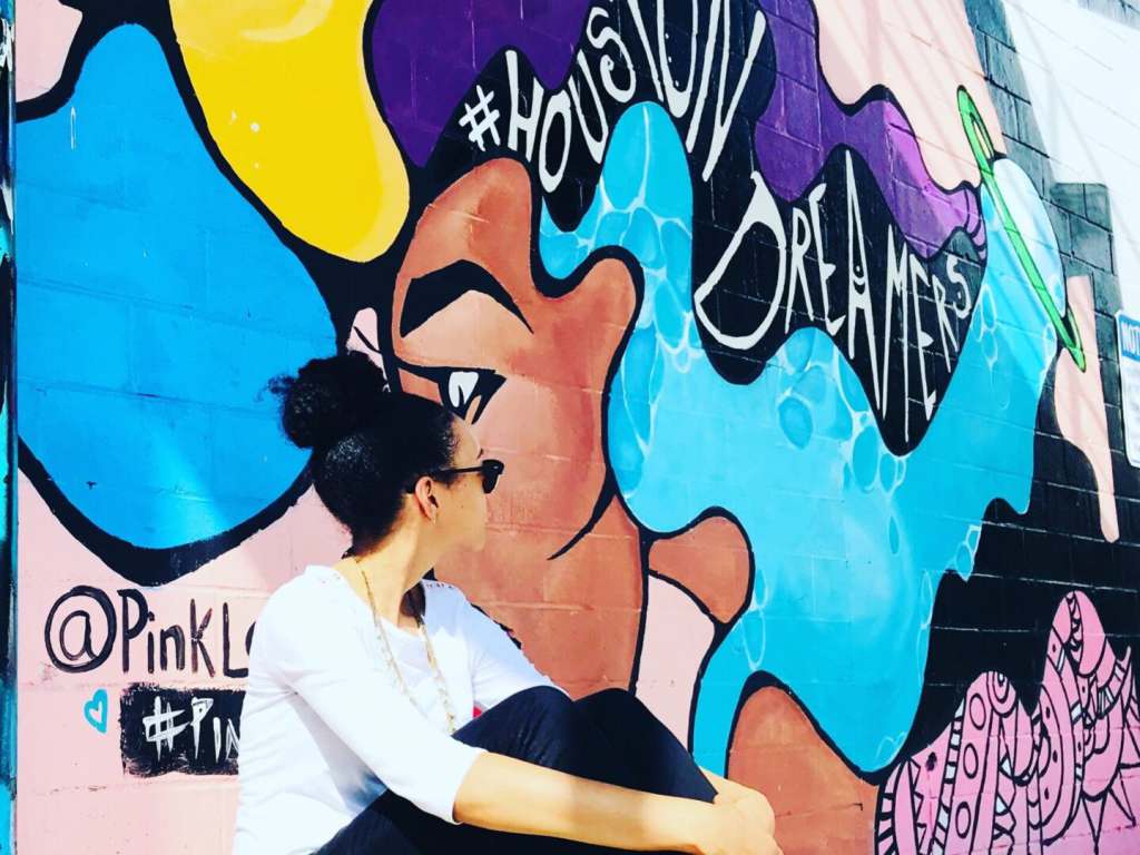 Cities with Incredible Black American Culture A TNAA employee sitting in front of a vibrant mural located on Leeland Street in Houston, Texas. The mural is created by an artist known as @PinkLomein.