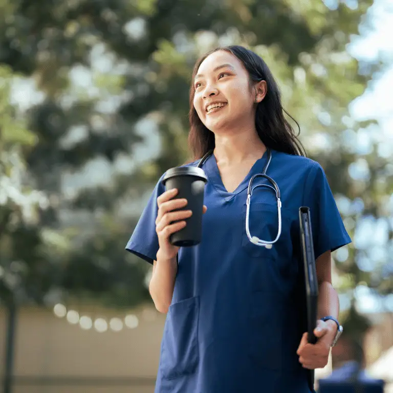 Healthcare professional in blue scrubs with a stethoscope walking outdoors while holding a coffee and tablet.