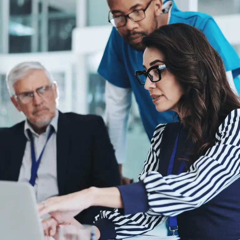 Healthcare leaders and clinical professionals collaborating around a laptop in a modern healthcare setting.