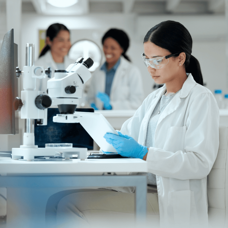Life sciences professional wearing safety glasses and gloves reviewing documents beside a laboratory microscope.