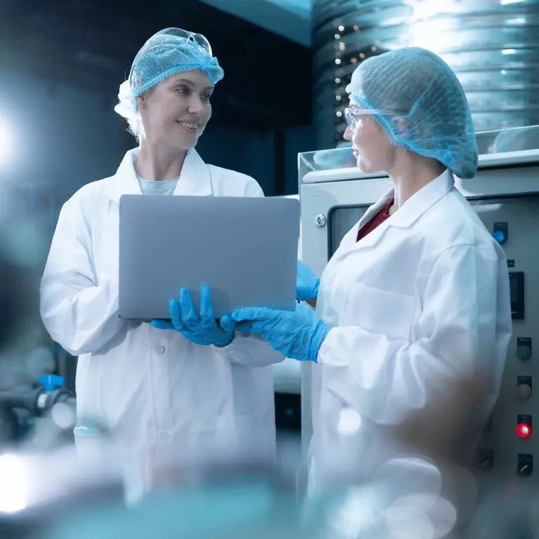 Two life sciences manufacturing professionals in protective equipment reviewing information on a laptop in a production facility.