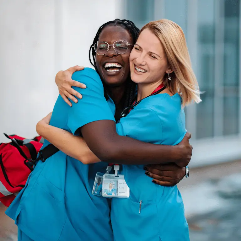 Two healthcare professionals in blue scrubs smiling and embracing outside a healthcare facility.