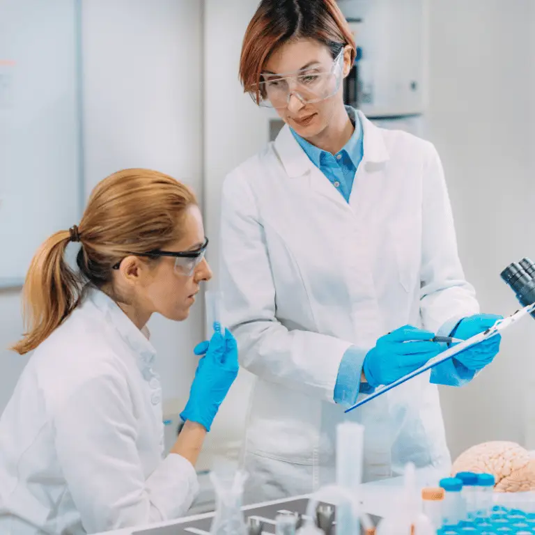 Two medical affairs professionals in lab coats and protective eyewear reviewing research findings together in a laboratory.